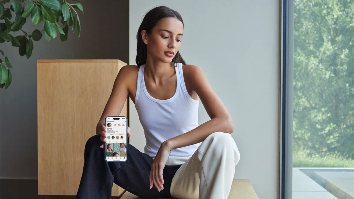 Modelo jovem sentada perto de uma janela grande num quarto claro, segurando o celular com o perfil do Instagram na tela mostrando uma grade de fotos, num momento tranquilo de ajuste do próprio perfil