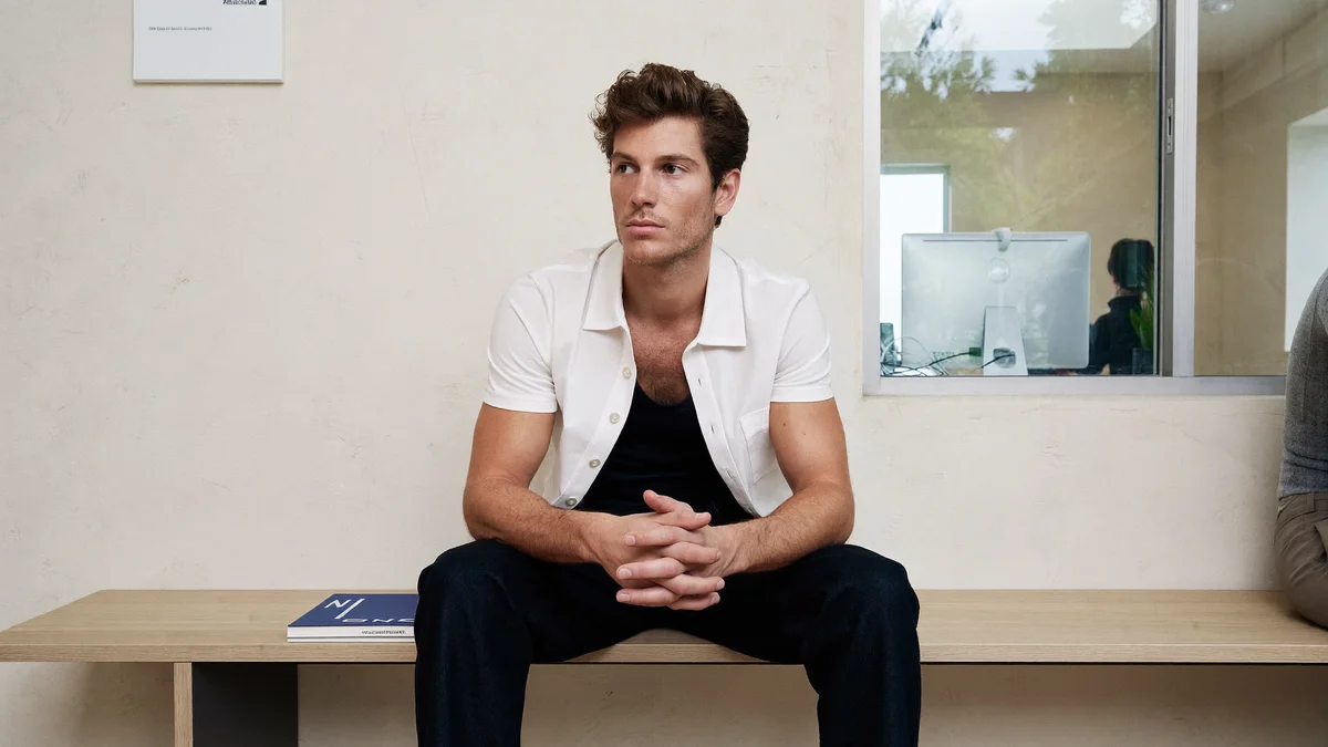 Young male model sitting on a wooden bench in the hallway of a modeling agency, white shirt open over a black tank top and dark trousers, hands clasped, looking to the side, a magazine on the bench and an office window with a computer in the background