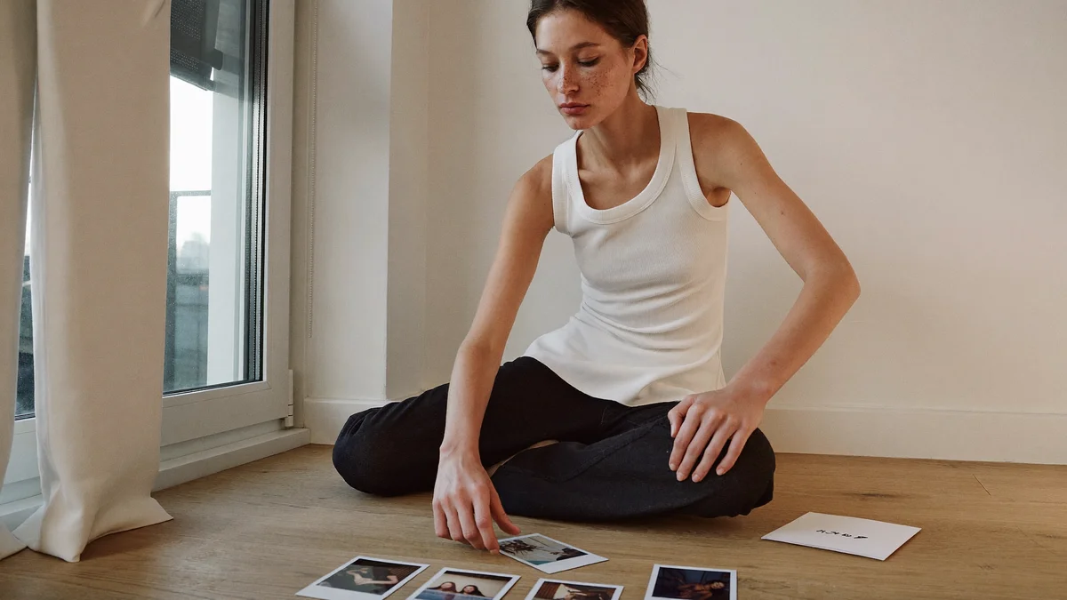 Modelo jovem sentada no chão de madeira de um apartamento claro, perto de uma janela grande, escolhendo entre polaroids impressas espalhadas à sua frente, num momento de preparo de material, sem posar pra câmera