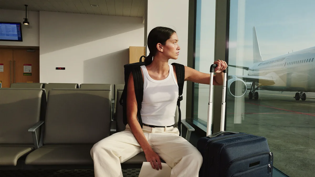 Modelo jovem sentada na sala de embarque de um aeroporto internacional, olhando pela janela grande um avião na pista na luz do fim de tarde, mochila nas costas e uma das mãos apoiada na alça da mala ao lado, num momento de espera antes da temporada