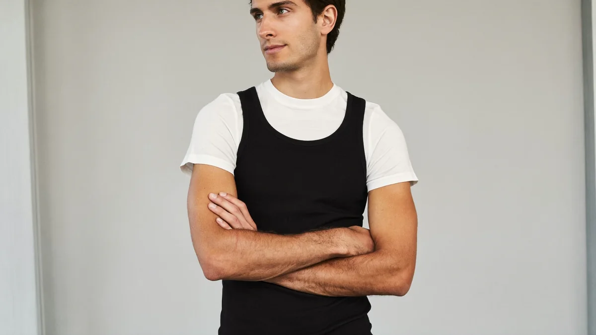 Young model with arms crossed in front of a light gray wall, white t-shirt under a black tank top, looking off to the side with a calm expression