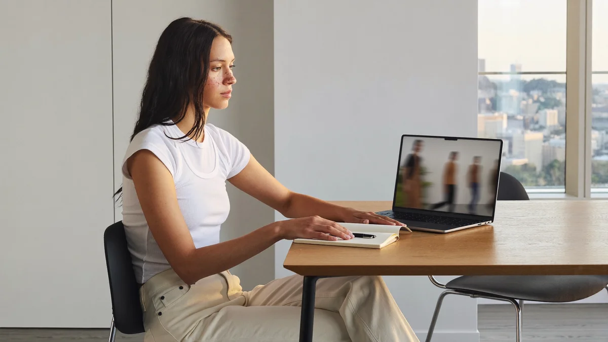 Modelo jovem assistindo a um desfile no laptop à mesa de madeira clara, caderno aberto ao lado, apartamento com skyline pela janela no fim da tarde