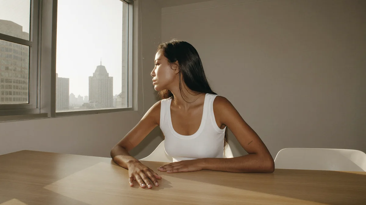 Young model seated at a table in a simple short-stay apartment, warm late-afternoon light, a large window behind her showing buildings of an Indian city in the distance, a closed book on the table with one hand resting on it