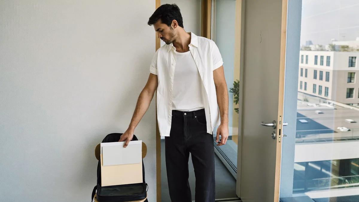Modelo jovem de manhã num apartamento compartilhado, guardando uma pasta lisa numa mochila aberta, pronto pra sair pra um dia de castings, prédios da cidade pela janela