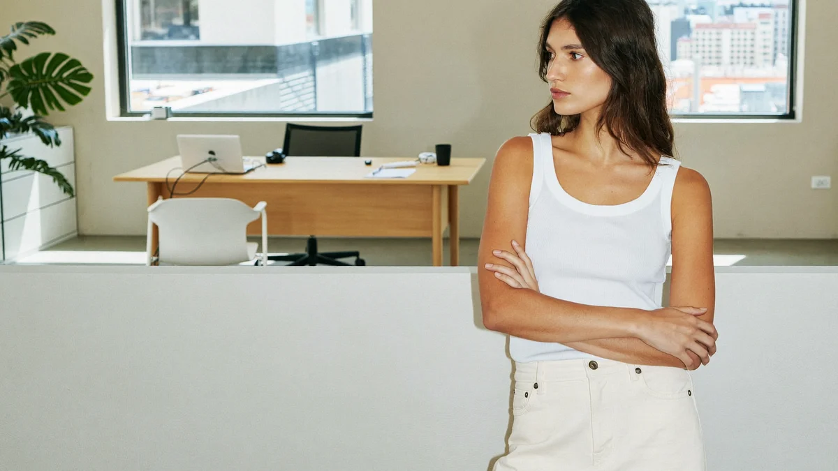 Young model standing in a model agency room, arms crossed, white tank top and cream trousers, looking to the side, with a desk, laptop, chair, plant and a window with a blurred city view behind her, daylight