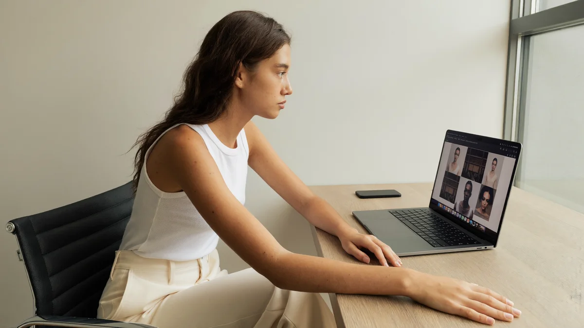 Modelo jovem numa mesa em casa, notebook aberto com uma grade de fotos na tela, luz de janela, revisando o próprio feed