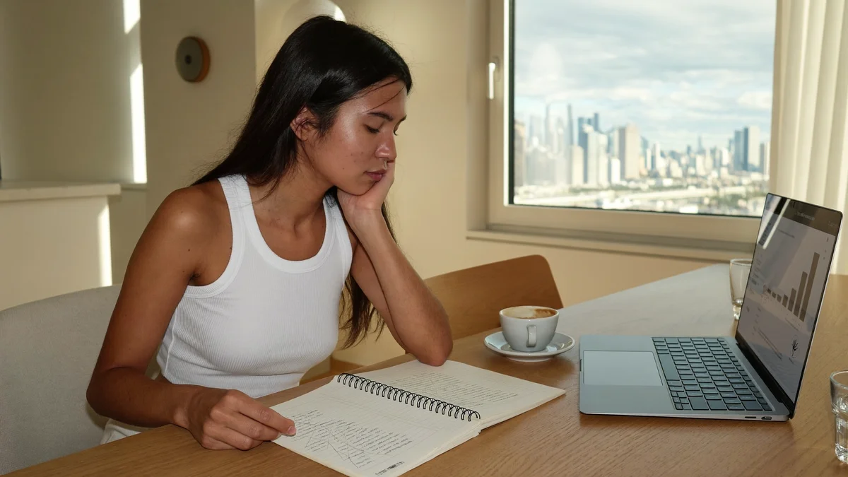 Modelo jovem fazendo as contas da temporada num caderno ao lado do laptop, apartamento claro com skyline pela janela no fim da tarde
