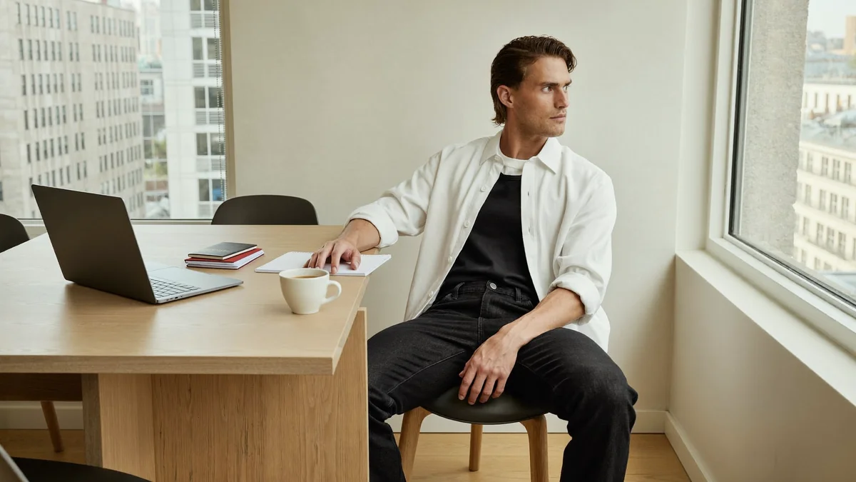 Modelo jovem sentado a uma mesa de madeira clara perto de uma janela grande num apartamento simples no exterior, laptop fechado ao lado, passaporte e caderno sobre a mesa, olhando a cidade lá fora num momento de pausa entre temporadas