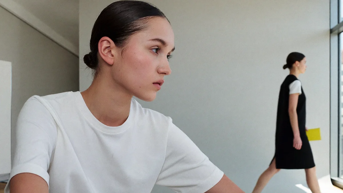 Model in a white T-shirt and low bun watching, from a bright casting studio, another model doing her walk in the background